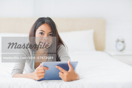 Young pretty girl lying on her bed using her tablet smiling at camera in her bedroom at home