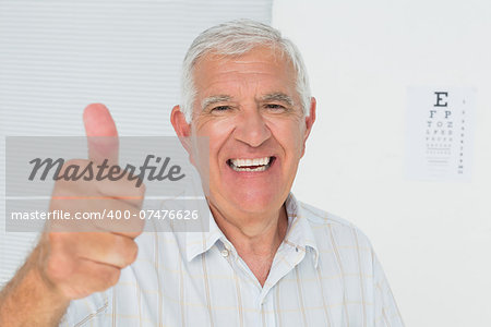 Portrait of a smiling senior man gesturing thumbs up with eye chart in the background at medical office