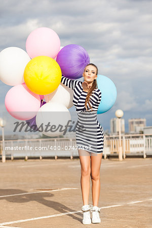 girl in black and white striped short dress and white high top sneakers sends air kiss holding bunch of multicolored balloons