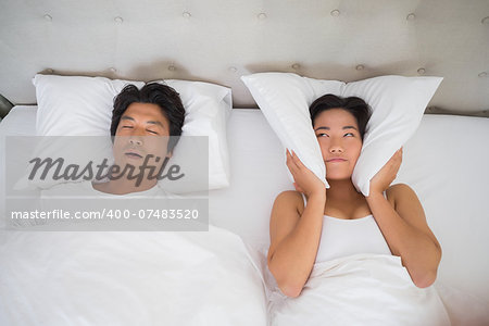 Annoyed woman covering her ears with pillows to block out snoring at home in bedroom