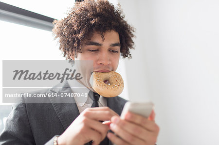 Young man using cell phone with bagel in mouth