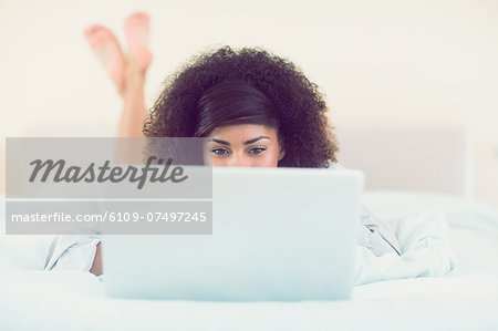 Pretty brunette lying on bed using laptop in bright bedroom