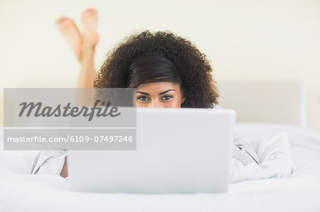 Pretty smiling  brunette lying on bed hidden behind laptop in bright bedroom