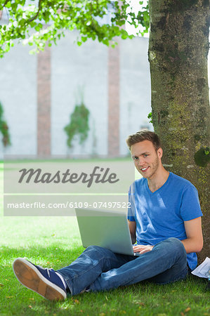 Cheerful handsome student sitting under tree using laptop on college campus