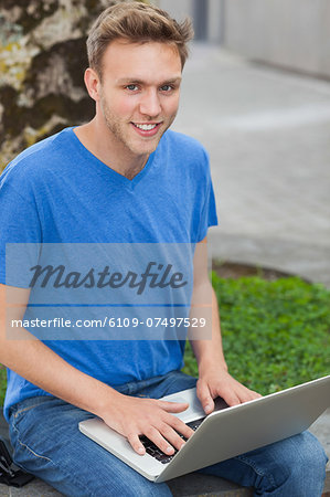 Cheerful handsome student sitting on wall using laptop on college campus