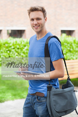 Happy handsome student holding tablet and standing on college campus
