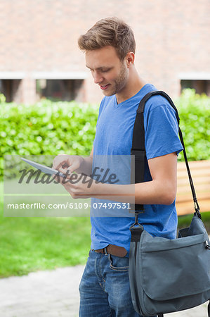 Happy handsome student using tablet and standing on college campus