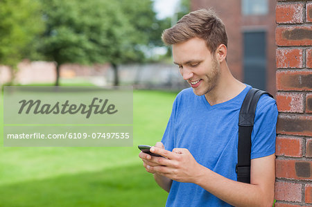 Smiling handsome student leaning against wall texting on college campus