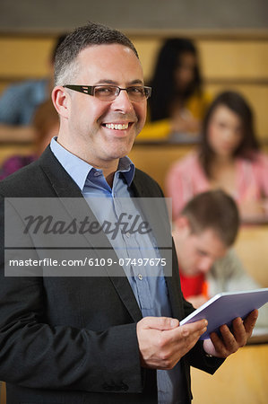 Lecturer holding tablet pc in front of class in lecture hall smiling at camera in college