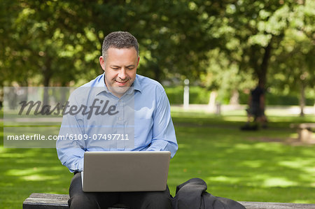 Cheerful lecturer sitting on bench using laptop on campus at the university