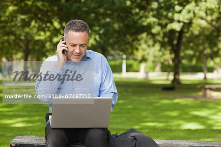 Cheerful professor talking on phone while sitting on bench using laptop on campus at the university