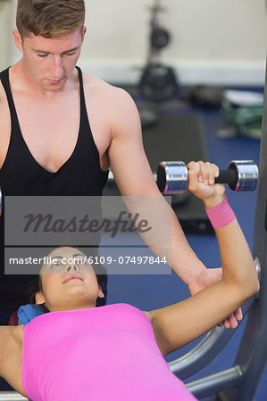 Instructor helping lying brunette exercising with dumbbells in weights room of gym