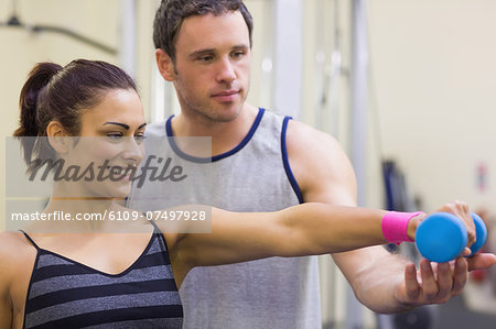 Instructor assisting cheerful woman lifting dumbbells in weights room of gym Instructor assisting cheerful woman lifting dumbbells in weights room of gym