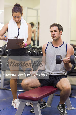 Trainer taking notes of muscular man lifting dumbbells in weights room of gym