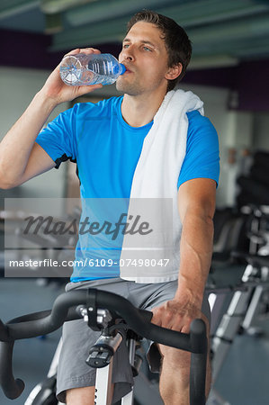 Tired young man drinking water while working out at spinning class in gym