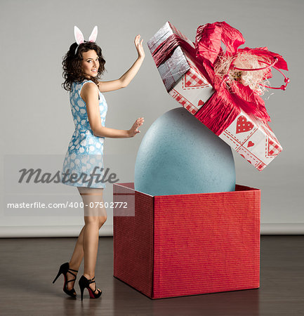 very cute young woman in spring ligh blue dress, bunny ears near big red box with easter egg inside