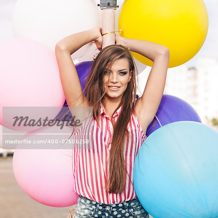 young smiling girl with messy hair in short denim shorts and sleeveless striped top leans on light-pole holding bunch of bright balloons