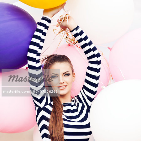 close-up of smiling girl with smokey eye make up, ponytail hair in short black and white striped dress keeps her hands raised holding bunch of multicolored balloons