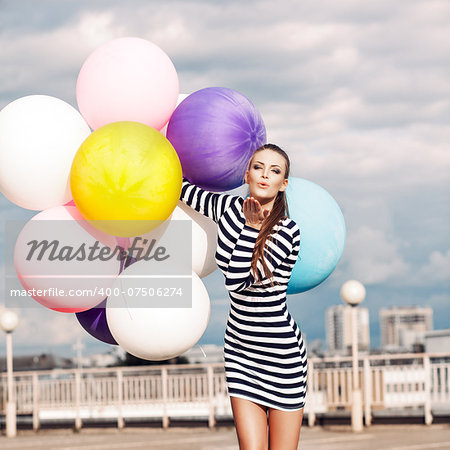 girl in black and white striped short dress and white high top sneakers sends air kiss holding bunch of multicolored balloons