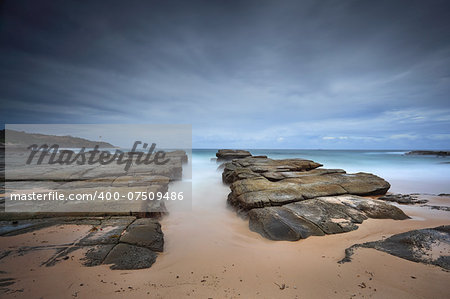 Stormy beach with foreground rocks at the edge of Soldiers point Norah Head NSW Australia.  Norah Head Lighthouse in distance. Long exposure