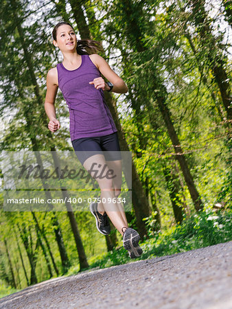 Photo of a young woman jogging and exercising on a gravel path through a forest. Slight motion blur on jogger.