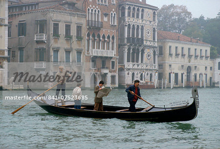 On a grey day in Venice a man commuting by gondola on the canal stands up to read his newspaper.