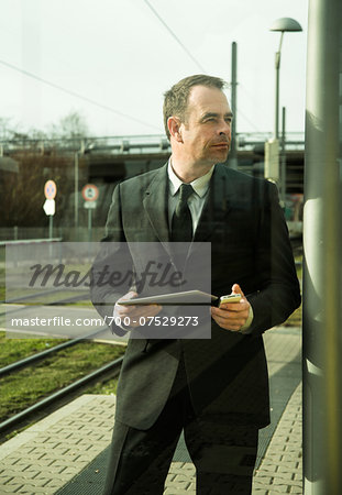 Portrait of businessman holding cell phone and tablet computer, standing at train station outdoors, Mannheim, Germany