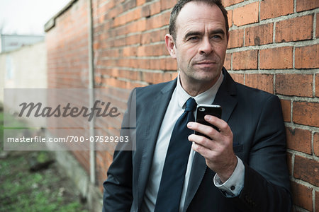 Portrait of businessman standing next to brick wall outdoors, holding cell phone, Germany