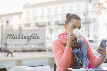 Young woman drinking coffee while using cell phone at sidewalk cafe