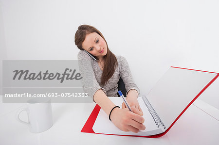 Businesswoman writing notes while answering smart phone in office