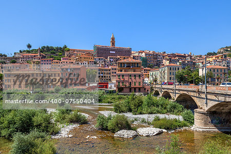 Urban bridge across riverbed overgrown with trees, shrubs and grass and old town of Ventimiglia under blue sky in Italy.