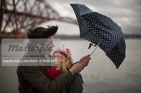 A young couple struggle with an umbrella in front of the Forth Rail Bridge in Queensferry, near Edinburgh, Scotland
