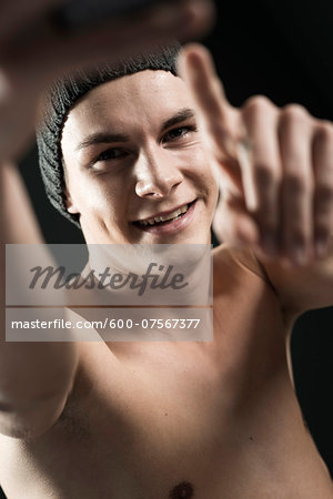 Close-up portrait of young man wearing toque, taking a selfie with cell phone, studio shot on black background