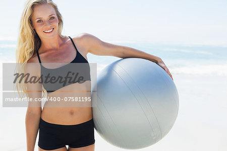 Fit blonde holding exercise ball at the beach smiling at camera on a sunny day