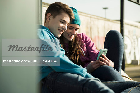 Teenage girl and boy sitting on ground, smiling and looking at cell phone together, Mannheim, Germany