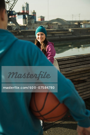 Teenage girl outdoors wearing toque, smiling and looking at teenage boy holding basketball, industrial area, Mannheim, Germany