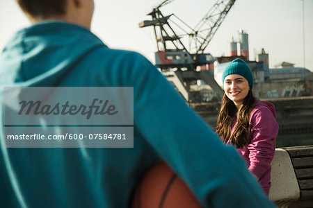 Teenage girl outdoors wearing toque, smiling and looking at teenage boy holding basketball, industrial area, Mannheim, Germany