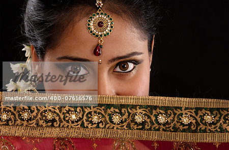 Portrait of beautiful mystery young Indian girl covering her face by veil, looking at camera, isolated on black background.