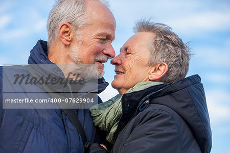 Attractive elderly couple in warm clothing standing clue together with outstretched arms, closed eyes and laughing smile against a blue sky embracing and celebrating the sun