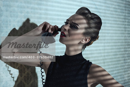 sensual elegant girl with creative hair-style and dark dress speaking on vintage phone in indoor half-light atmosphere with interrogative expression