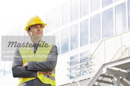 Young male architect in protective wear standing arms crossed outside building