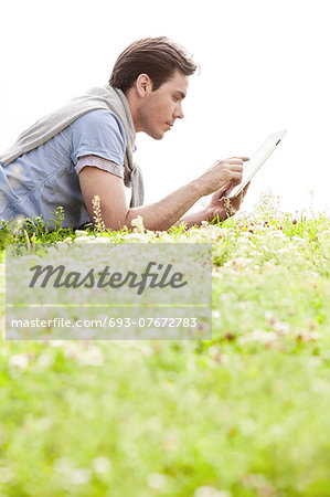 Side view of young man using digital tablet while lying on grass against clear sky