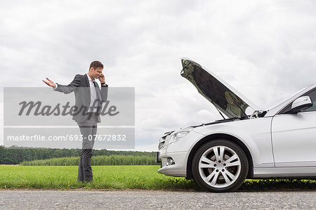 Frustrated young businessman using cell phone by broken-down car at countryside
