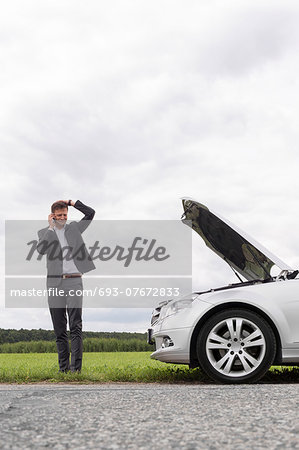 Frustrated young businessman using cell phone near broken down car at countryside