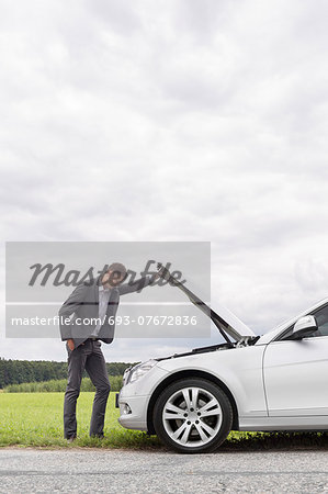 Side view of young businessman examining broken down car engine at countryside
