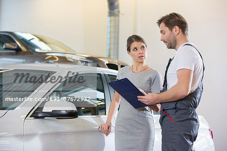 Repairman holding clipboard while conversing with female customer in automobile repair shop