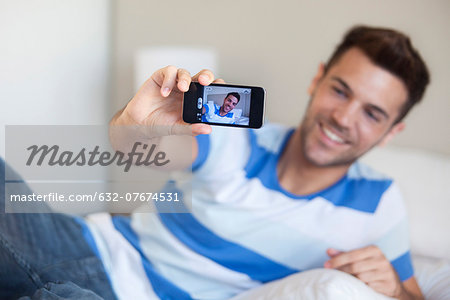 Young man reclining on bed taking selfie with smartphone