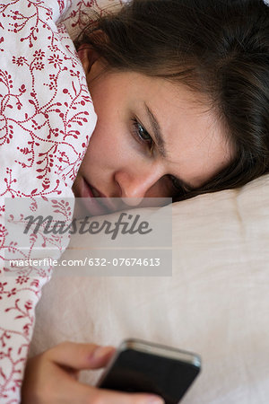 Woman lying bed looking at smartphone with unhappy expression on face