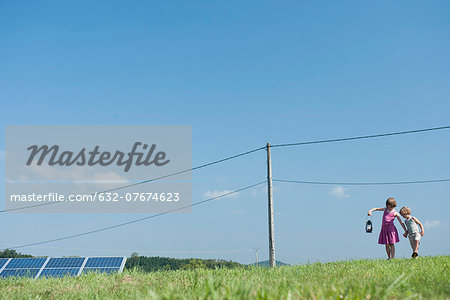 Children walking in field near solar panels and power lines, carrying old-fashioned lantern