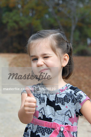 Little girl gives a "thumbs up" sign and winks.  She is playing outside and is enjoying the great outdoors.